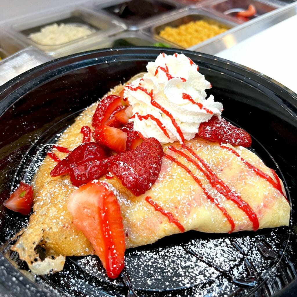 A crepe with strawberries, strawberry syrup, whipped cream and powdered sugar on top.