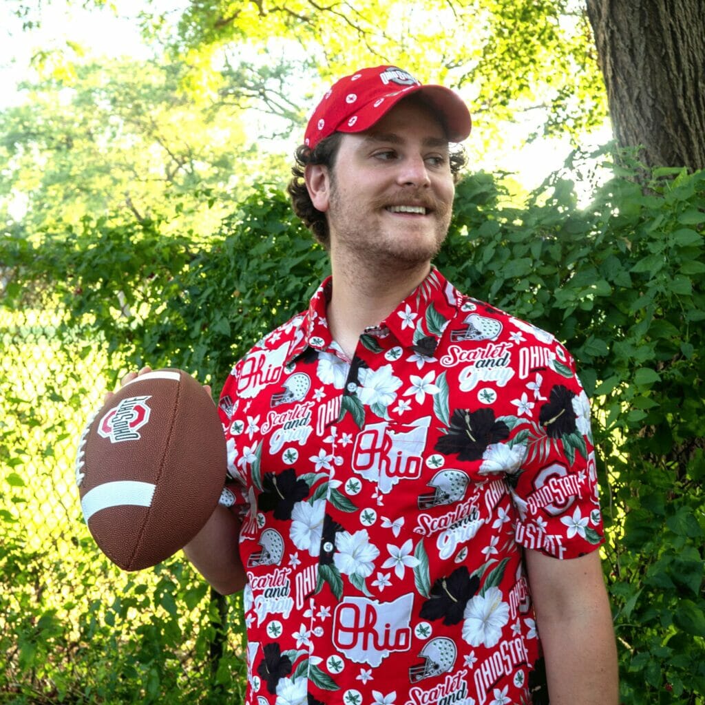 A man wearing Ohio State Buckeye Gear and standing outside holding a football.
