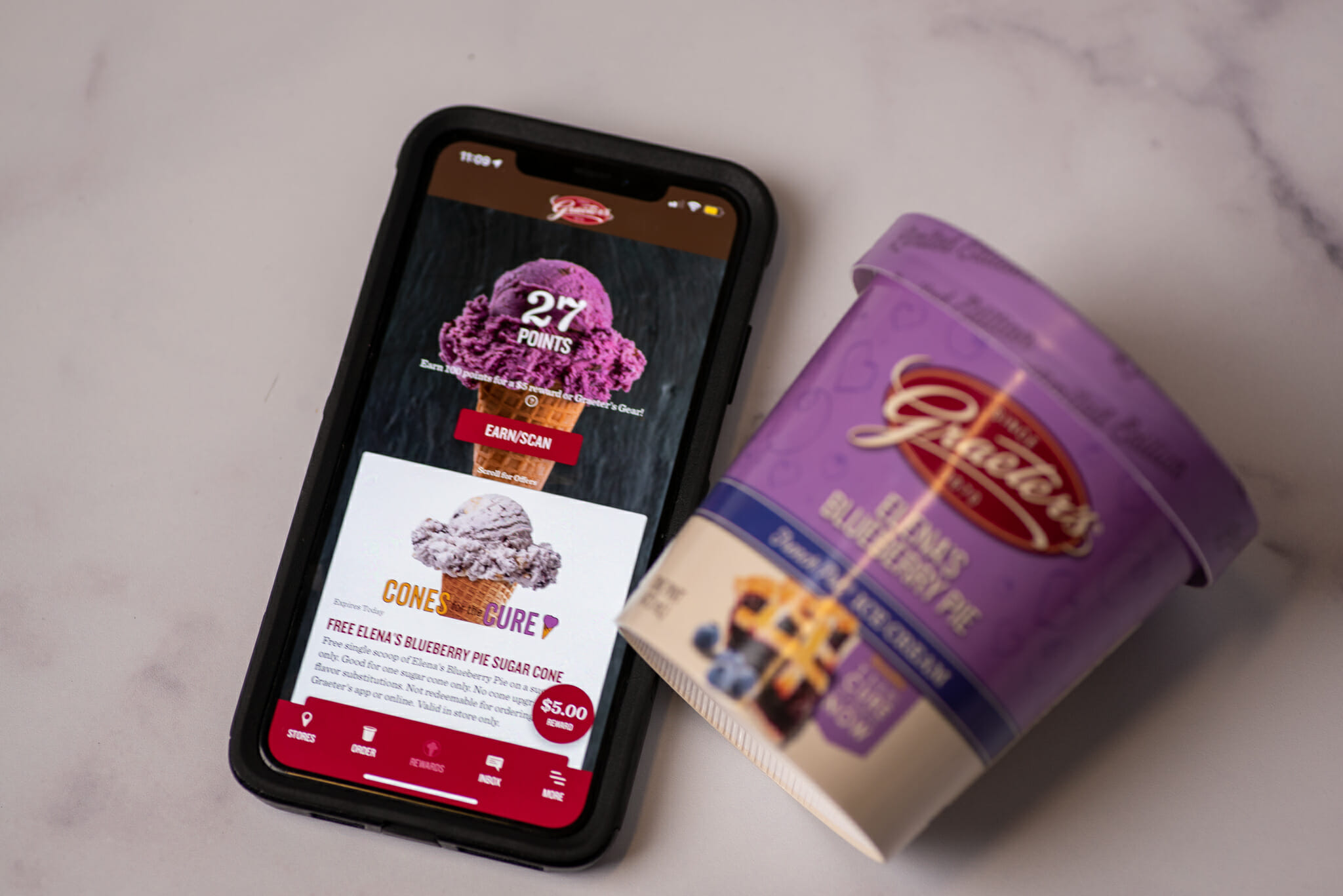 An photo of a cell phone with the Graeter's app pulled up and displaying the Cones for the Cure fundraising information. Next to the cell phone is a small container or Graeter's ice cream.