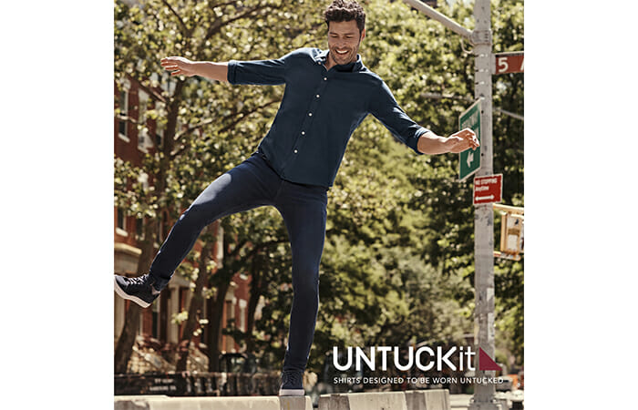 A man balancing on a sidewalk barricade wearing UNTUCKit clothing with promotional copy that reads UNTUCKit. Shirts designed to be worn untucked.