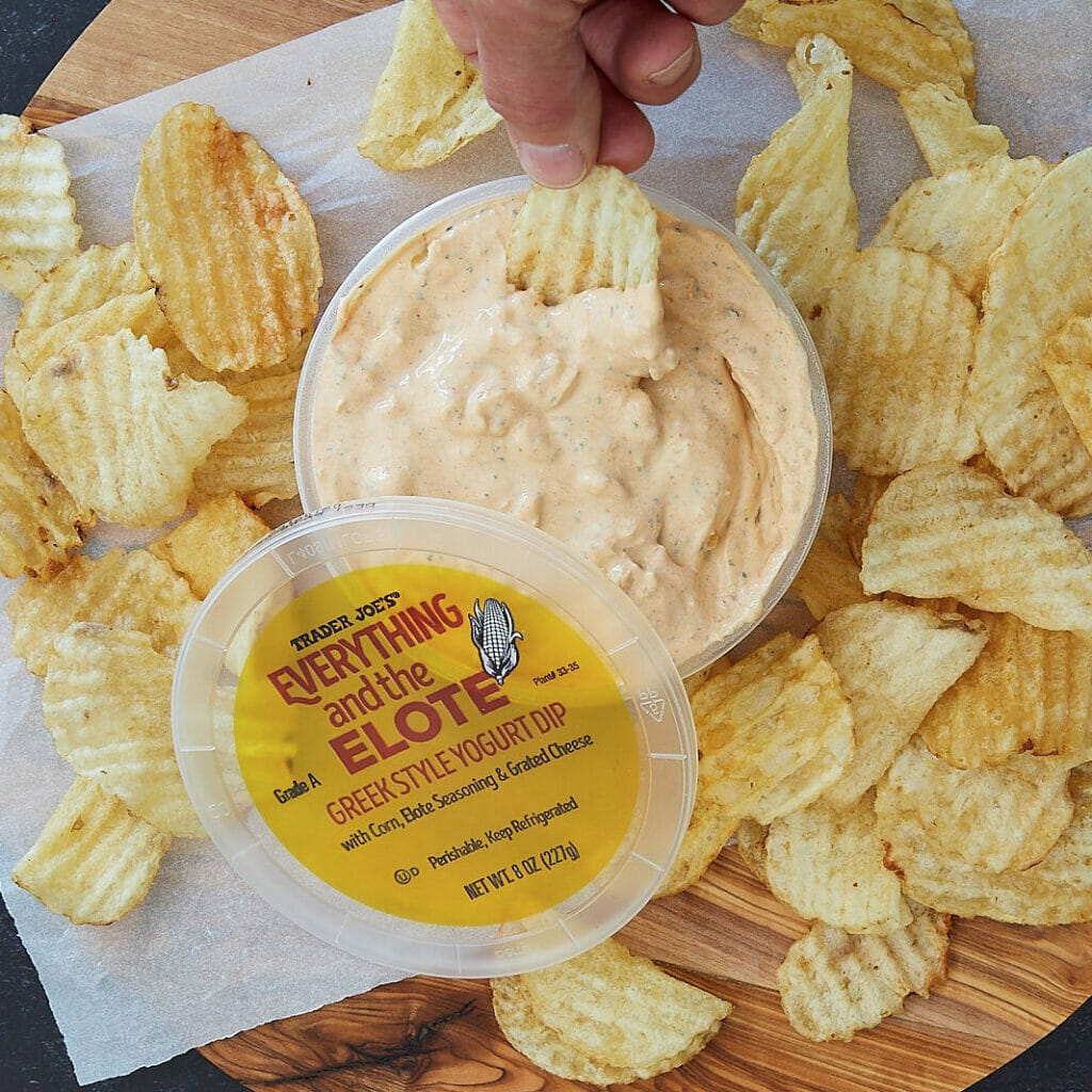 An overhead view of chips on a table with a container of dip in the middle and someone's hand can be seen about to dip a chip into the dip container. 
