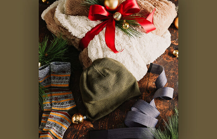 A blanket with holiday ribbon around it next to a beanie hat and a pair of comfy socks.