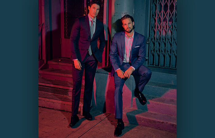 Two men sitting against a building wearing Indochino suits.