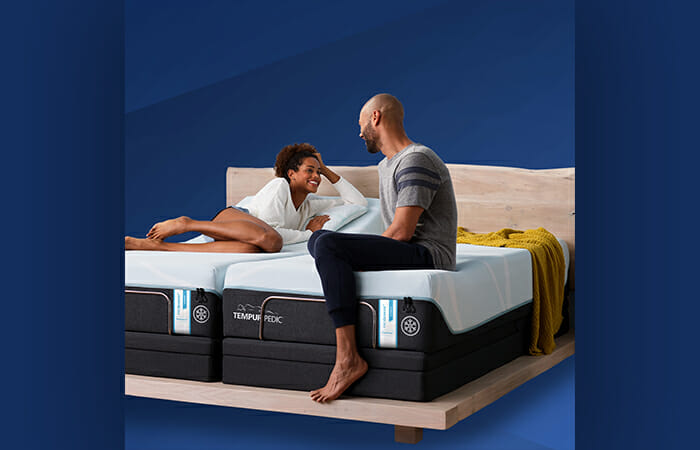 A man and a women sitting on a Tempur-pedic bed.