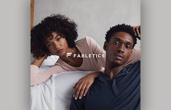 A man and a woman leaning on a bed wearing Fabletics clothing.