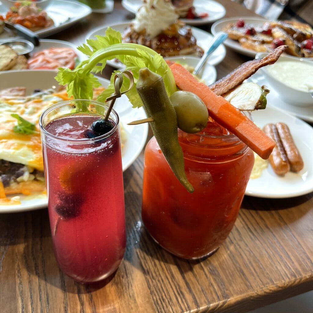 A mimosa and a bloody mary in glasses on a table with garnishes and plates of food in the background.