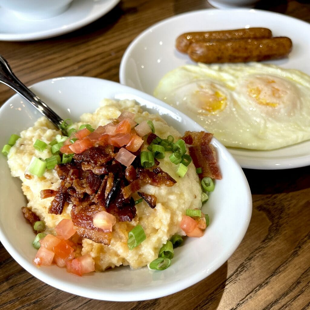 Two breakfast meals from Another Broken Egg at Easton Town Center consisting of savory grits with bacon, tomato and chives on top and a plate of two eggs with a side of sausage links next to it on a table. 