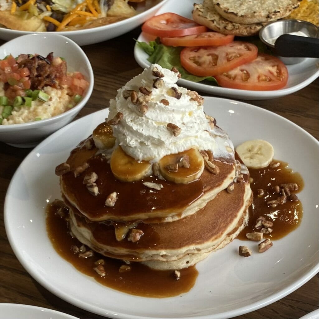 A breakfast meal from Another Broken Egg at Easton Town Center consisting of banana pancakes with bananas, whipped cream and pecans on top. Plates of food can be seen in the background behind the pancakes as well.