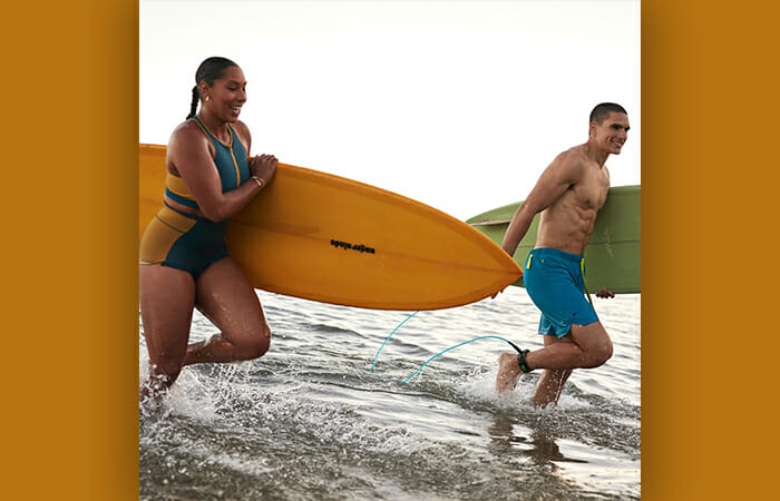 A man and a woman holding surf boards and running through shallow water while wearing Fabletics swim gear.