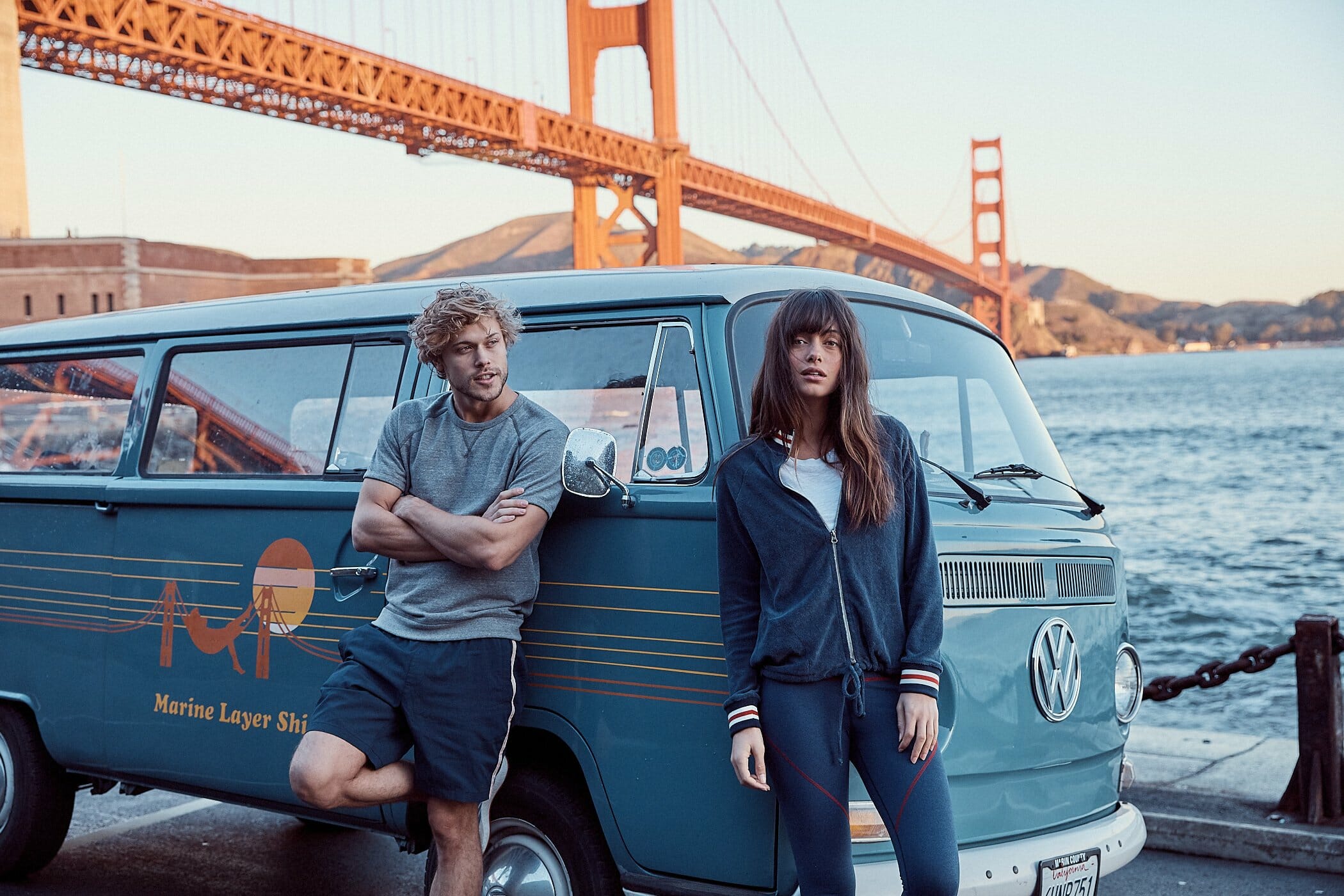 A man and a woman wearing Marine Layer clothing and satding in front of an old VW van in front of the GOlden Gate Bridge.