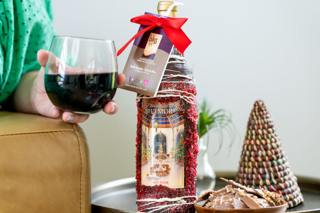 bliss in a bottle wine bottle, glass of wine, and pieces of chocolate sitting next to a small christmas tree.