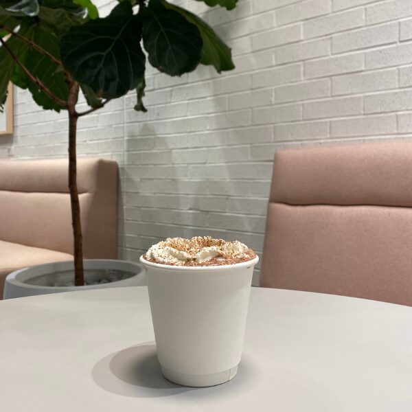 A cup of hot chocolate from Capital One Cafe sitting on a table with a plant and two pink benches visible in the background.