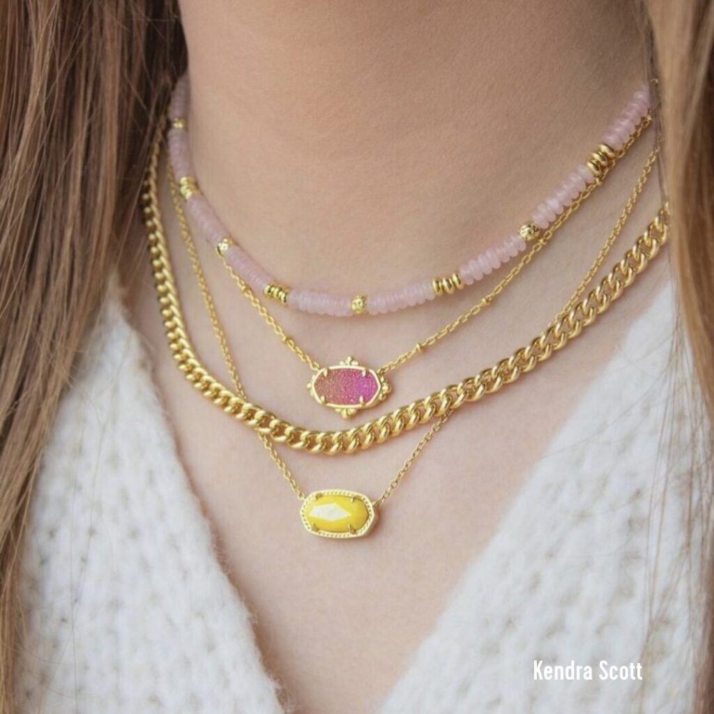 A close up of a woman's neck line with 5 necklaces layered from Kendra Scott.
