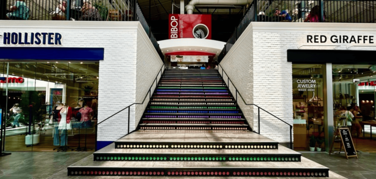 The Hollister and Red Giraffe Designs storefronts inside the Station Building at Easton with the Grand Staircase between them lit up with rainbow colors.