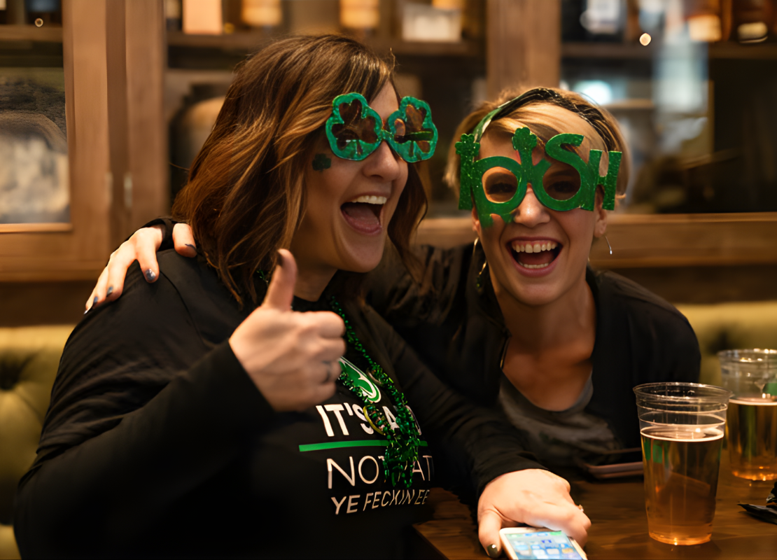 Two women smiling and laughing inside a pub wearing shamrock sunglasses.