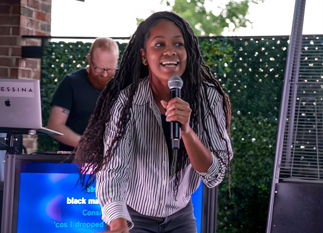 Women smiling and holding a microphone singing karaoke outdoors with a DJ standing in the background.