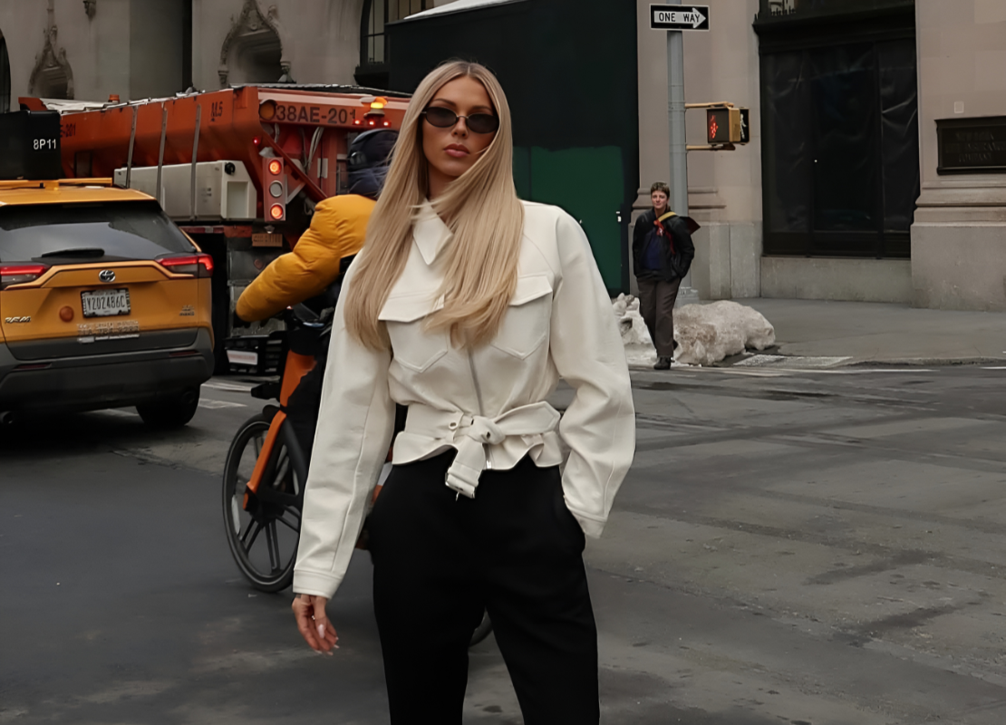 Women with long blonde hair wearing round sunglasses, white jacket and black pants standing in the street.