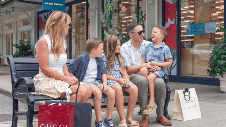 A family sitting on a bench in front of the Warby Parker store at Easton with a child sitting on the dad's lap and laughing. Shopping bags are on the ground beside them. A mother, a boy, and a girl watch as the dad and son laugh.