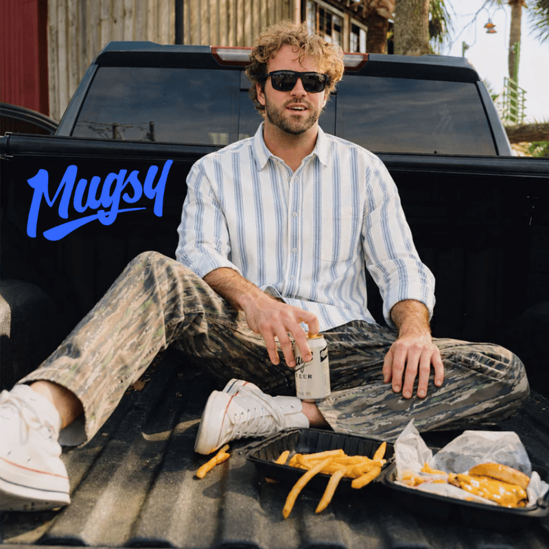 a man sitting in the bed of a truck with a beer and french fries wearing Mugsy clothing.